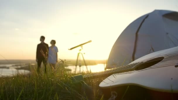 夕暮れ時に屋外夏休みを楽しんでいる若いカップルの前に芝生の上のギター — ストック動画