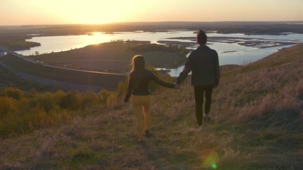 Ungt par promenader i ett fält som håller händerna vid solnedgången — Stockvideo