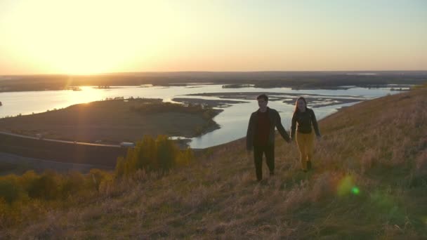 Jovem casal atravessa a colina de mãos dadas ao pôr do sol — Vídeo de Stock