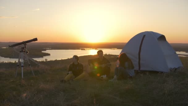 Grupa przyjaciół, siedząc na trawie w pobliżu namiotu chłodzenie o zachodzie słońca — Wideo stockowe