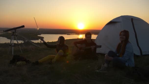 Glada unga vänner sitter på gräset och ha roligt tillsammans i solnedgången utomhus — Stockvideo