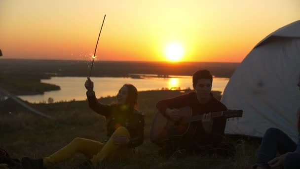 Glada unga vänner att ha roligt tillsammans och spelar gitarr i solnedgången på kullen — Stockvideo