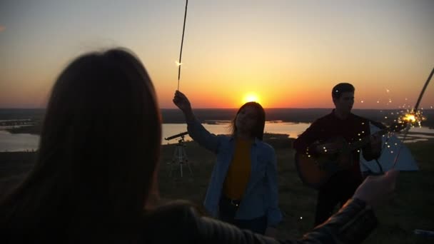 Fiatal nők tánc csillagszórók mögöttük srác gitározni a dombon napnyugtakor — Stock videók