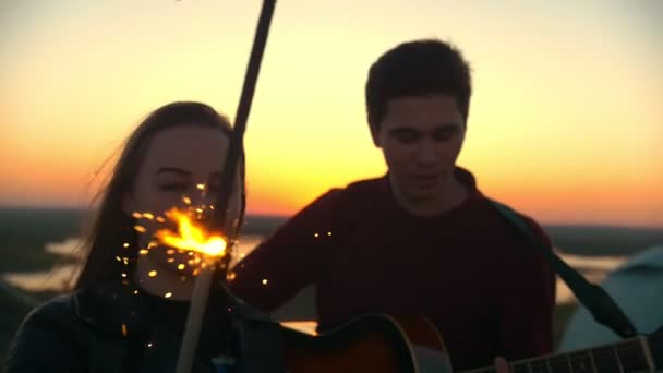 Mujer joven bailando con sparkler delante de su novio tocando la guitarra al aire libre al atardecer — Vídeos de Stock