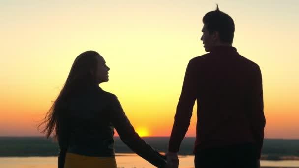 Visão traseira casal de mãos dadas na colina olhando uns aos outros no pôr do sol incrível — Vídeo de Stock