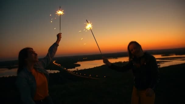 Two happy young women dancing with sparklers on a hill at summer sunset — Stock Video