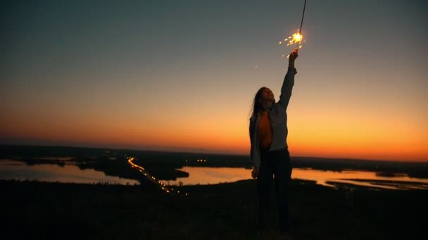 Jovem mulher segurando sparkler na mão ao pôr do sol incrível na colina — Vídeo de Stock