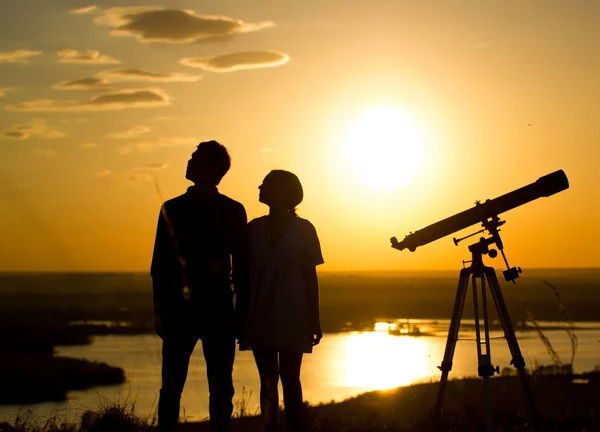 Siluetter av par - pojke och flicka nära teleskopet på kullen på sommar solnedgång — Stockfoto