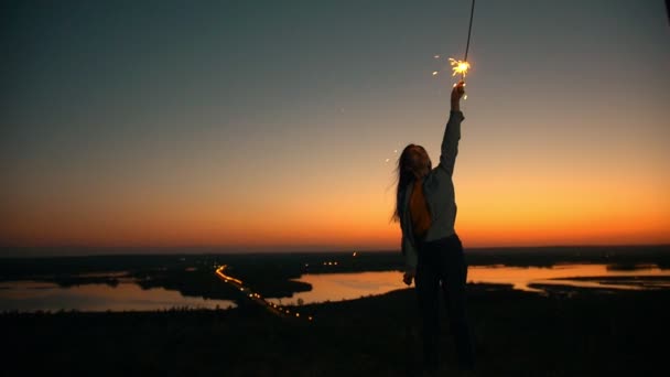 Glad ung kvinna med långt hår som flyger i vinden höja handen med tomtebloss på sommar solnedgång på kullen — Stockvideo
