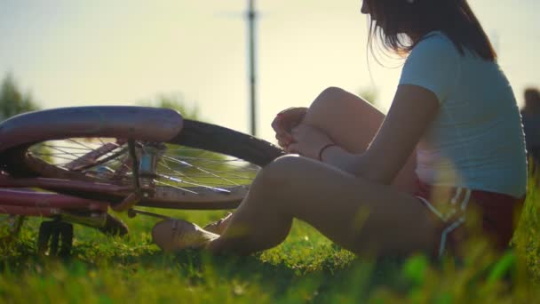 Meisje zittend op het gras en schuift het wiel van een fiets, op de achtergrond rijden een ander meisje, zonnige dag — Stockvideo