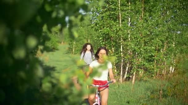 Branches d'arbres verts, filles riant et se rattrapant sur les vélos, arbre vert, journée d'été ensoleillée — Video