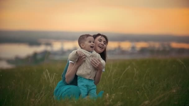 Дівчина сидить на зеленій траві поруч з нею її дитина, вони сміються, захід літнього дня — стокове відео