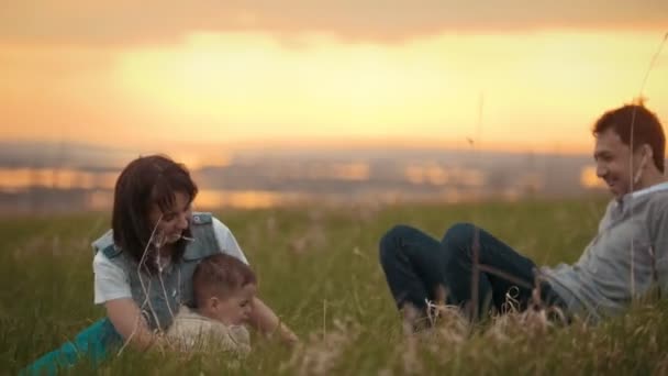 Πατέρα και μητέρα που παίζουν με το γιο της, το μωρό κάθονται στο γρασίδι με ένα παιχνίδι, καλοκαιρινό βράδυ στη φύση — Αρχείο Βίντεο