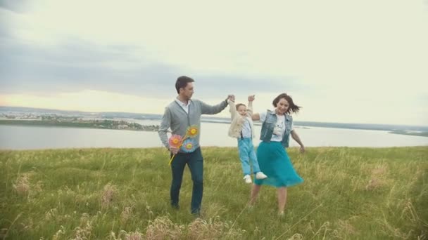 Семья идет через поле на фоне реки, мама и папа держат ребенка за руки, облачно — стоковое видео