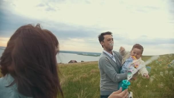 Малыш наслаждается мыльными пузырьками в руках отца, ходит по полю в облачный летний день — стоковое видео