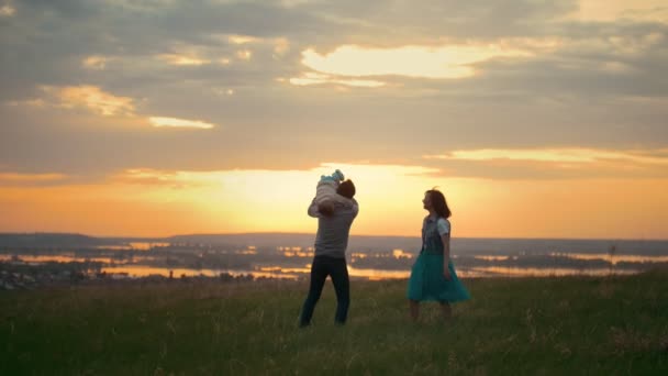 Jovens pais brincam felizes com seu filho, a mãe corre com o bebê, um pôr do sol brilhante, uma noite de verão. — Vídeo de Stock