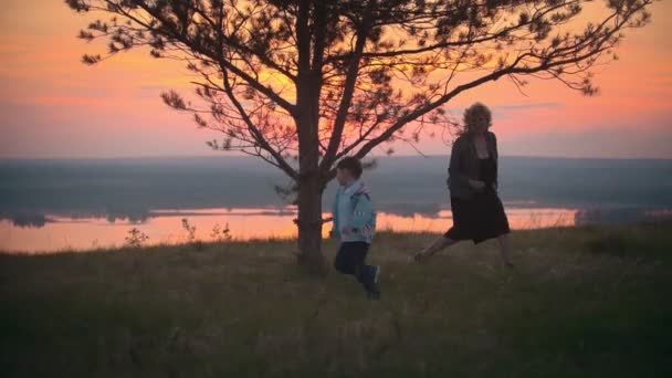 Oma en kleinzoon bij zomer zonsondergang spelen en rennen de boom oma probeert te vangen een kleinzoon, ze lachen dat ze plezier hebben — Stockvideo
