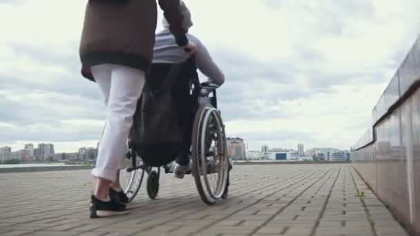 Nő hordozza egy fogyatékkal élő ember egy tolószékben, az utcán — Stock videók