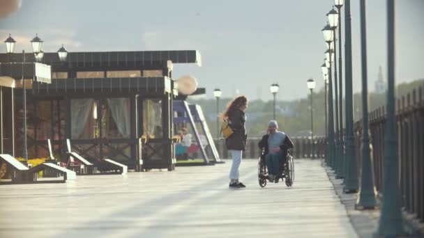 Инвалид в инвалидном кресле разговаривает по телефону, гуляя вместе со своей девушкой на набережной. — стоковое видео
