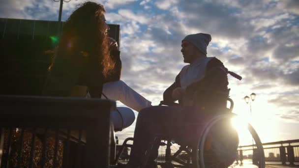 Jovem com homem deficiente em uma cadeira de rodas falando ao ar livre ao pôr do sol — Vídeo de Stock