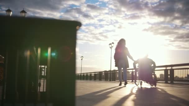 Jonge vrouw met gehandicapte man in een rolstoel naar de kant van de Bank — Stockvideo