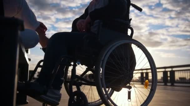 Sziluettjét a fiatal nő, fogyatékossággal élő ember egy tolószékben, beszél a naplemente szabadban — Stock videók