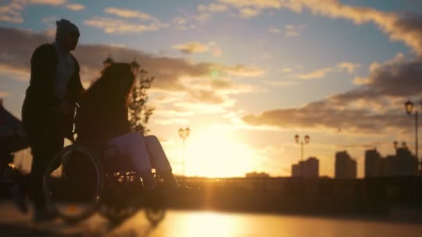 Caring man with a disabled woman in wheelchair walking through the quay at sunset — Stock Video