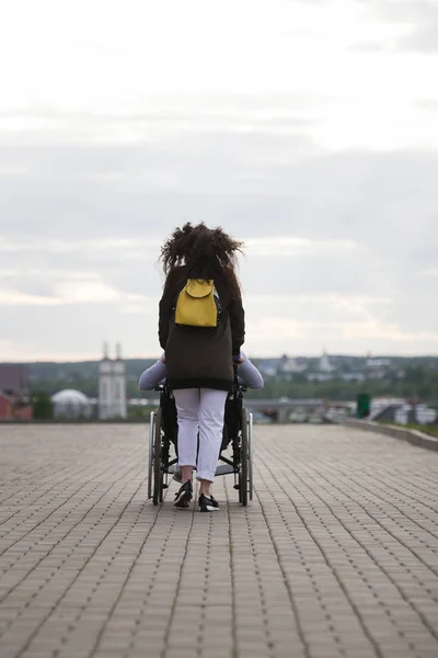 Rückansicht einer jungen Frau, die mit einem behinderten Mann im Rollstuhl am Kai spazieren geht — Stockfoto