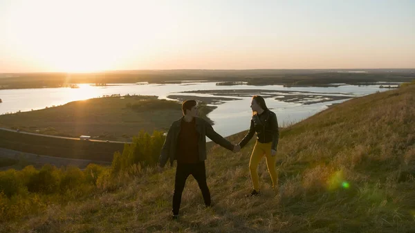 Молода пара проходить через пагорб, тримаючи руки на заході сонця — стокове фото