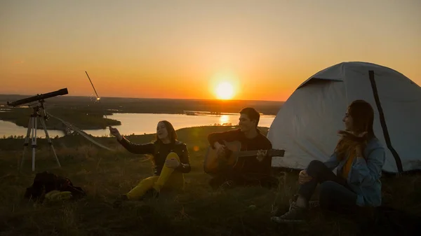 Alegre jóvenes amigos divirtiéndose juntos y tocando la guitarra al atardecer en la colina —  Fotos de Stock