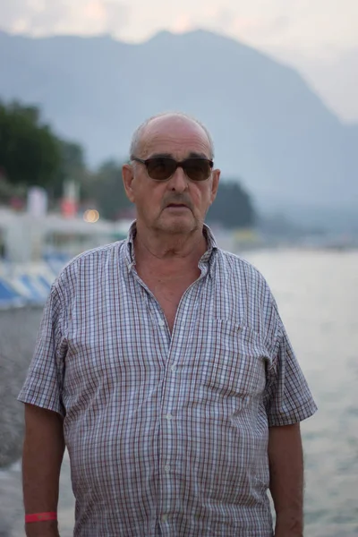 Porträt eines älteren Mannes mit Sonnenbrille am Meer vor dem Hintergrund der Berge — Stockfoto
