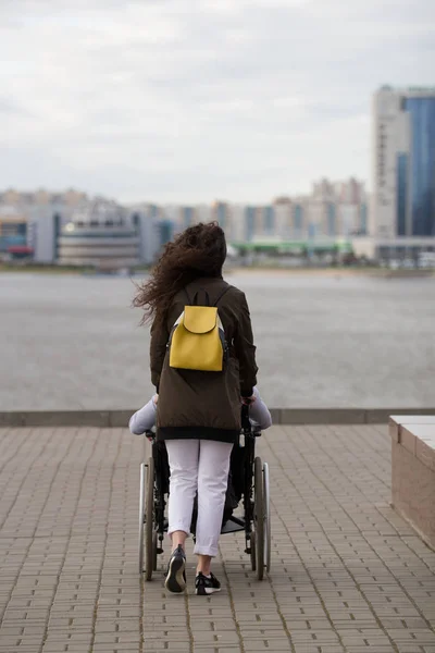 Rückansicht einer jungen Frau, die mit einem behinderten Mann im Rollstuhl am Kai spazieren geht — Stockfoto