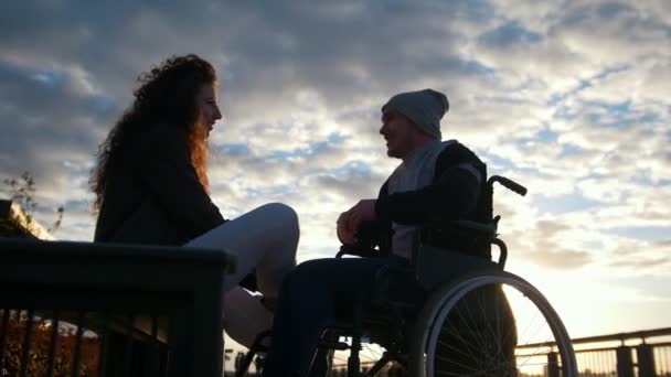 Pareja feliz - joven discapacitado en silla de ruedas con una joven disfrutando de la puesta de sol juntos — Vídeos de Stock