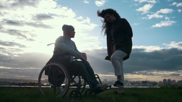 Soins aux handicapés - homme en fauteuil roulant avec une femme parlant au coucher du soleil nuageux — Video