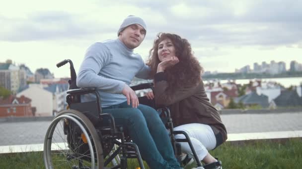 Ritratto di un uomo disabile felice in sedia a rotelle abbraccia una giovane donna all'aperto — Video Stock