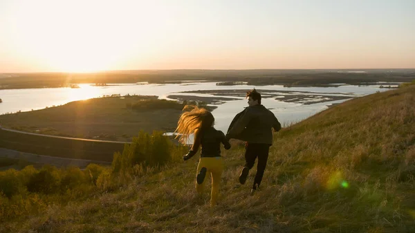 Щаслива молода пара біжить на пагорбі тримаючи руки на заході сонця — стокове фото