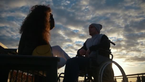Fiatal nő, fogyatékossággal élő ember egy tolószékben beszél szabadban naplementekor — Stock videók