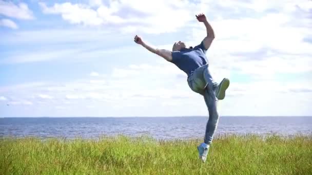 Молодой человек выполняет акробатические трюки на холме на фоне реки — стоковое видео