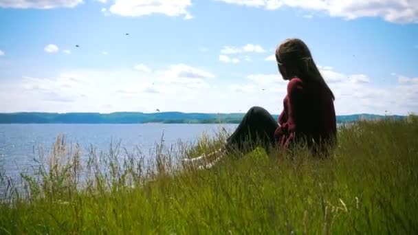 Mujer joven pensativa sentada en la colina mirando el río en un día soleado — Vídeos de Stock