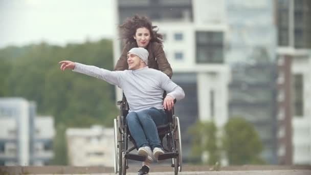 Happy gehandicapte man in een rolstoel met gelukkige jonge vrouw lopen bij de stad straat — Stockvideo