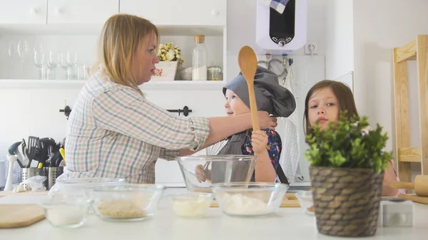 Mom puts a hat on her little girl who holding a wooden spoon in hand and elder girl looking at camera in a cooking studio