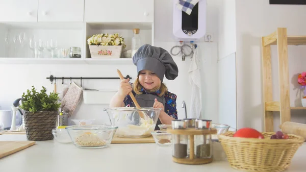 Счастливая маленькая девочка шеф-повар смешивает тесто с большой деревянной ложкой в стеклянную миску — стоковое фото