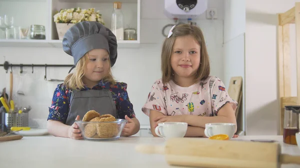 Две маленькие сестры улыбаются с домашним печеньем и чашками чая в кулинарной студии — стоковое фото