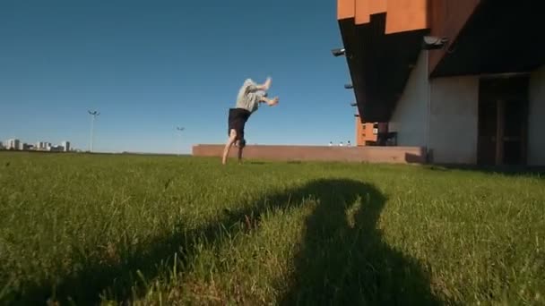 Jovem acrobático masculino executa um flip na grama verde ao sol — Vídeo de Stock