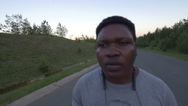 Joven afroamericano caminando por el camino vacío y fumando cigarrillos al atardecer — Vídeos de Stock