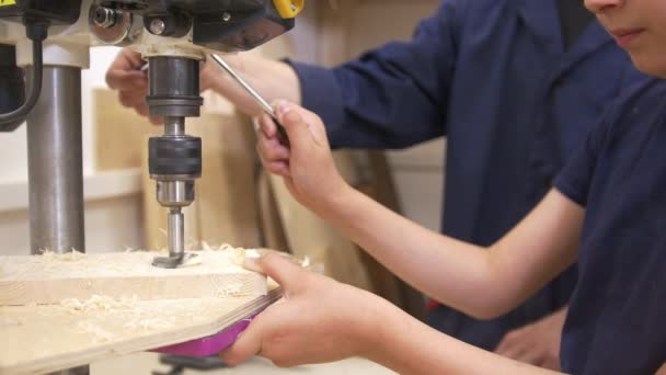 Adolescente chico perfora un agujero en un tablón de madera utilizando equipo industrial en taller de carpintería — Vídeos de Stock