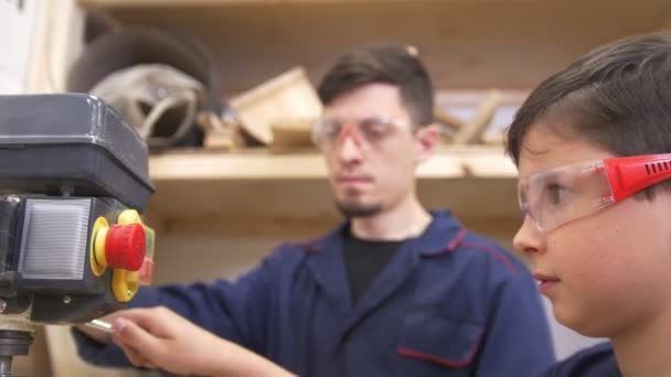 Adolescente niño con su padre taladra la tabla de madera utilizando la máquina de carpintería industrial en el taller — Vídeo de stock