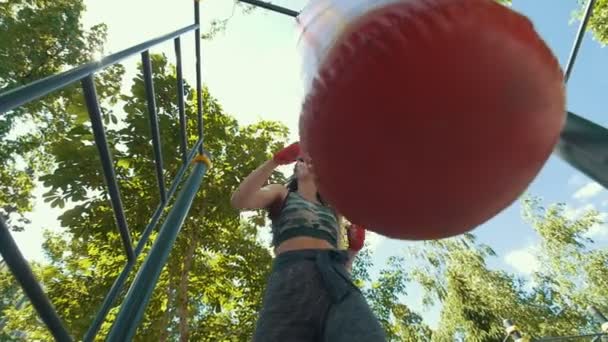 Атлетичная молодая женщина в спортивной одежде бьет боксерскую грушу, тренировка в летнем парке - вид снизу, замедленная съемка — стоковое видео