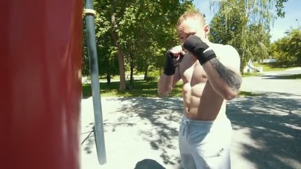 Atlético jovem envolvido em boxe com saco de perfuração no parque de verão — Vídeo de Stock