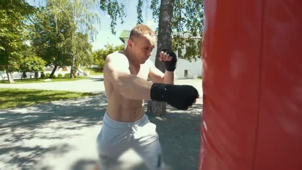 Muscular jovem envolvido em boxe com saco de perfuração no parque de verão — Vídeo de Stock